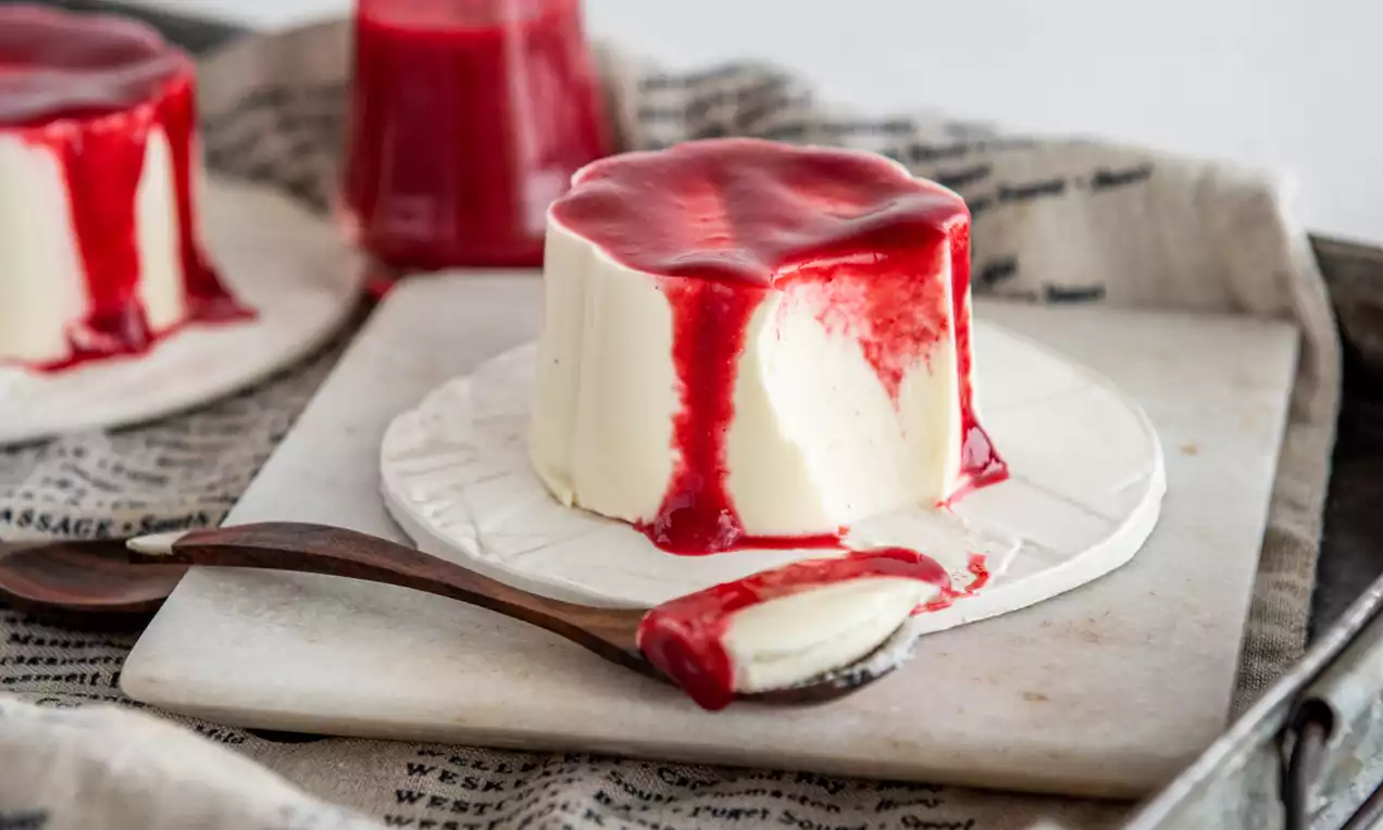 Picture - Vanille panna cotta met gladde, ivoorwitte textuur en helderrode saus, geserveerd op een wit bord met een houten lepel in een metalen tray