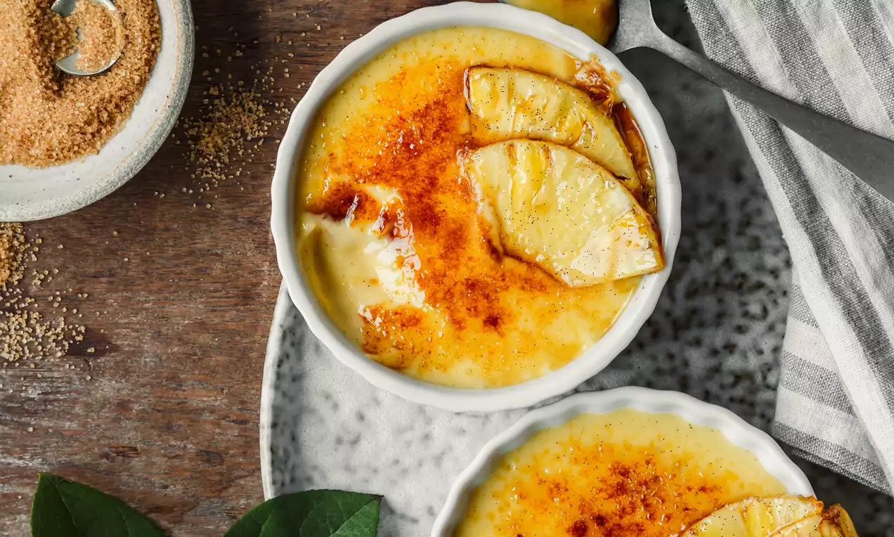 Picture - Coconut crème brûlée met goudbruine gekarameliseerde suikerlaag, gegarneerd met ananasschijfjes in twee witte bakjes op een gevlekt bord