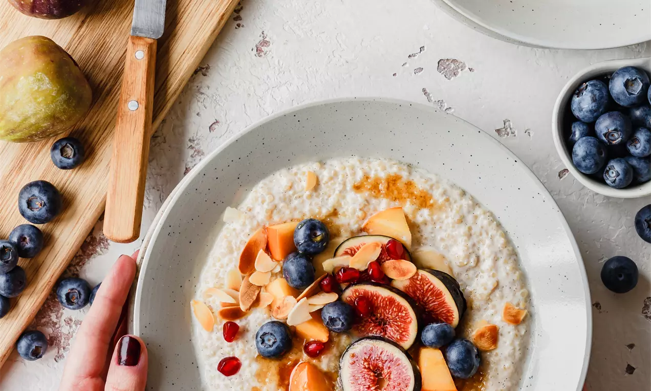 Picture - Quinoa vanillepap in een grijs diep bord gegarneerd met vijgen, blauwe bessen, pompoenblokjes en amandelschaafsel op een lichte ondergrond