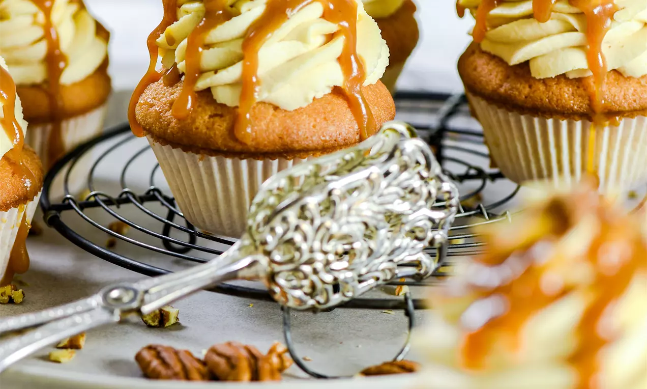 Picture - Cupcakes met lichtbruine cakebodems, opgespoten roomwitte crème, gegarneerd met stroperige karamel en stukjes pecannoten op een metalen rooster