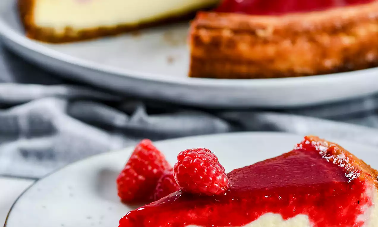 Picture - Driehoekige punt van een cheesecake met een gladde rode frambozentopping op een stevige bruine korst, gegarneerd met verse frambozen op een wit bord