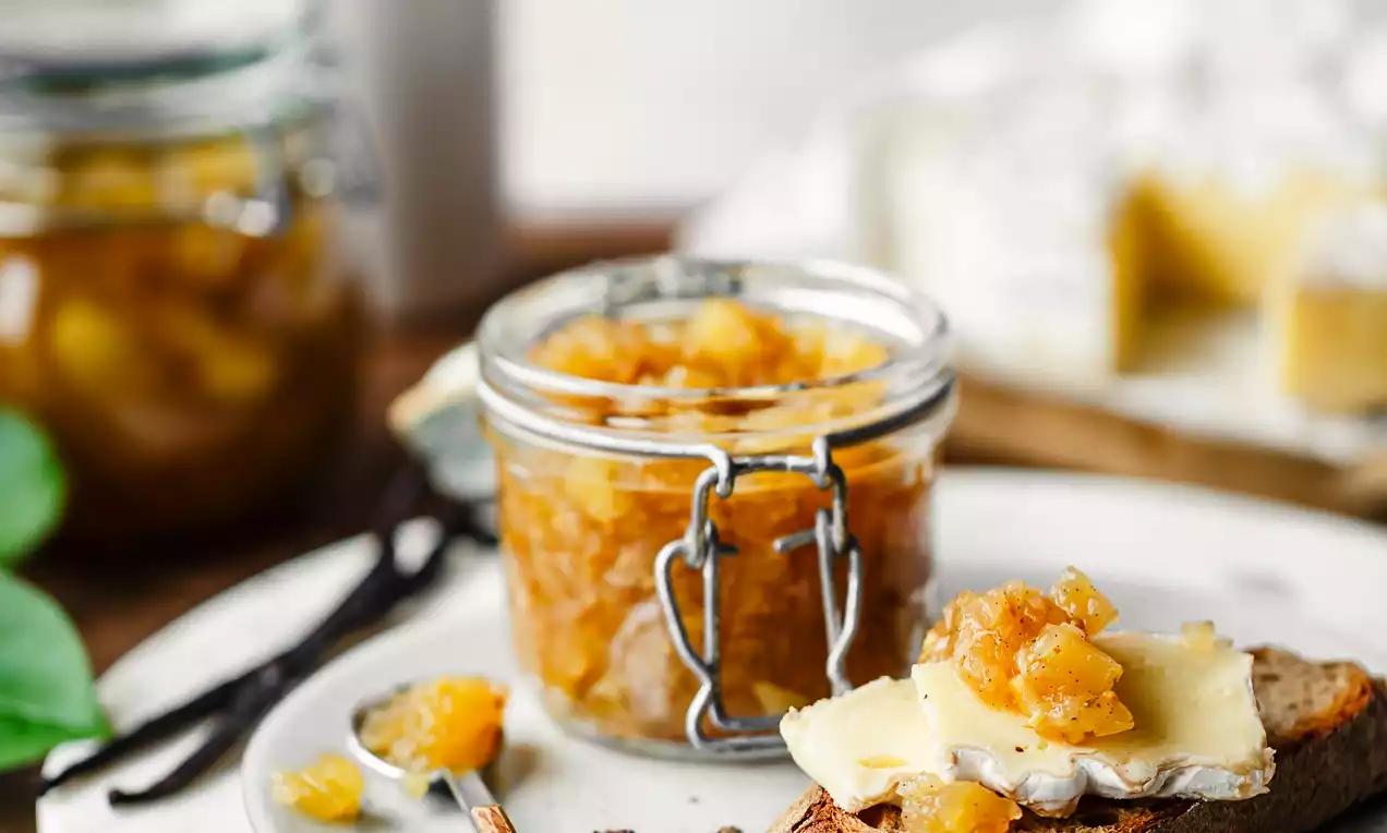 Picture - Op een houten tafel een pot met goudgele appelchutney, een snee brood met chutney en kaas, op een wit bord met kaneelstokje en kruidnagels