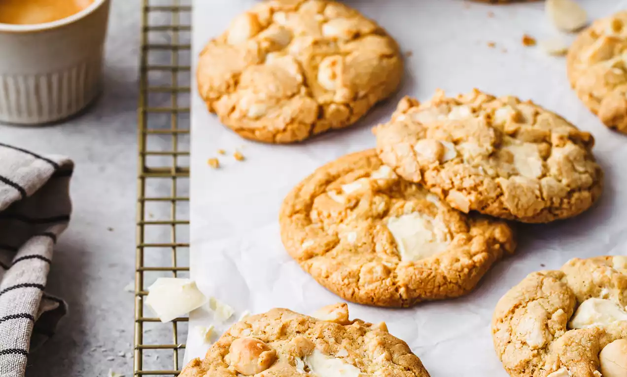 Picture - Goudbruine, ronde cookies met stukjes witte chocolade en macadamianootjes op bakpapier op een metalen rooster, omringd door schaaltjes en koffie