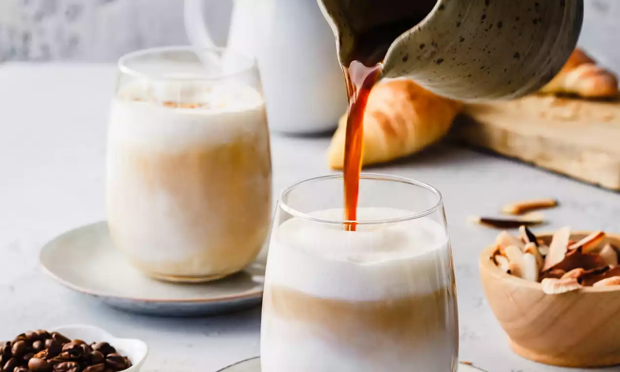 Picture - Coconut vanilla latte met zachte witte schuimlagen en lichtbruine tonen in glazen op een bord, omringd door koffiebonen en amandelschilfers